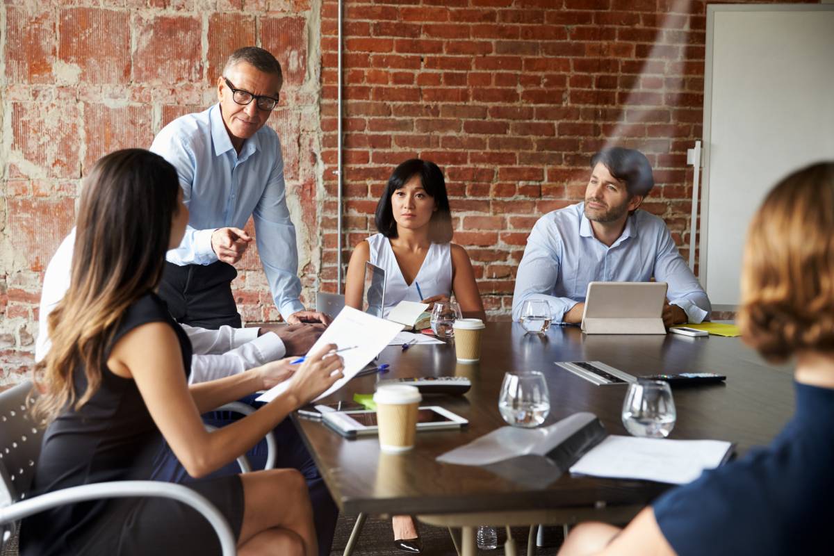 A diverse group of professionals in a meeting around a table with laptops and documents in a room with a brick wall